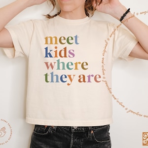 May include: A white cropped t-shirt with the text "meet kids where they are" in colorful letters. The text is in a playful font and is centered on the shirt.