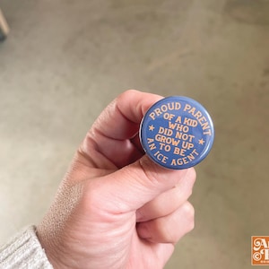 May include: A blue button with white text that reads "PROUD PARENT OF A KID WHO DID NOT GROW UP TO BE AN ICE AGENT." The button is held in a hand. The Angie Pea logo is in the bottom right corner.