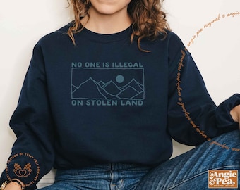 May include: Navy blue sweatshirt with the text "NO ONE IS ILLEGAL ON STOLEN LAND" in a teal color. The design includes a mountain range and a moon. The sweatshirt has long sleeves and a crew neck.