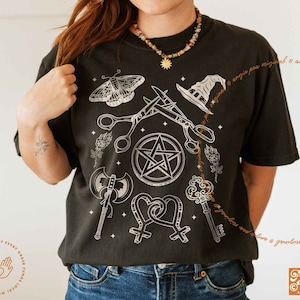 May include: Black t-shirt with a white graphic design featuring a pentagram, scissors, a witch's hat, a butterfly, an axe, a key, and a heart with a venus symbol. The text "Angie Pea. Original Design" is at the bottom of the shirt.