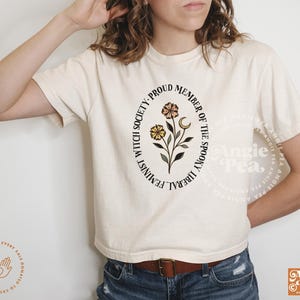 May include: A white t-shirt with a black circle graphic that says "Feminist Witch Society - Proud Member of the Spooky Liberal"