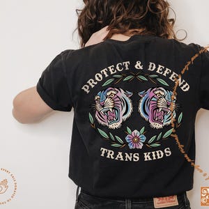 Pu&ograve; includere: Maglietta nera con una grafica di due tigri colorate con il testo "Protect & Defend Trans Kids" stampato sul retro.