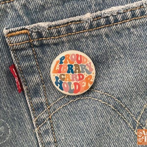 May include: A round button with the text "PROUD LIBRARY CARD HOLDER" in a retro font, featuring orange, blue, and brown colors. The button is set against a backdrop of blue denim jeans.