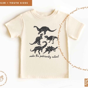 May include: A cream colored t-shirt with a black graphic of five dinosaurs and the text "make the patriarchy extinct".