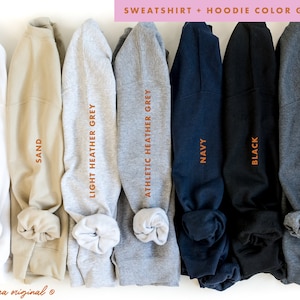 May include: A color guide for sweatshirts and hoodies, showcasing a range of colors. The image displays folded sweatshirts in white, sand, light heather grey, athletic heather grey, navy, black, and dark heather grey. Text on the image reads "SWEATSHIRT + HOODIE COLOR GUIDE."
