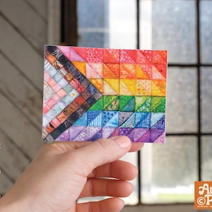 May include: A square sticker featuring a colorful geometric design resembling a quilt or patchwork pattern. The design incorporates a rainbow of colors and patterns. The sticker is held in a hand, with the Angie Pea logo visible.