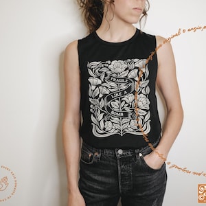 May include: Black sleeveless top with a white floral design and the text "FRAGILE LIKE A BOMB" on a banner. The top is paired with black jeans. The Angie Pea original design logo is visible.
