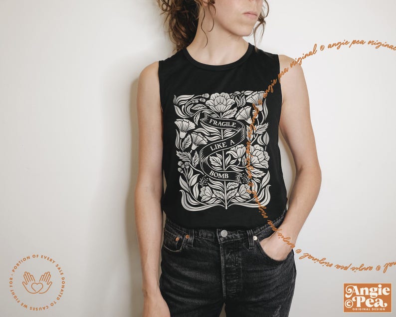 May include: Black sleeveless top with a white floral design and the text "FRAGILE LIKE A BOMB" on a banner. The top is paired with black jeans. The Angie Pea original design logo is visible.