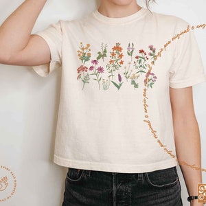 May include: A white cropped t-shirt with a floral design featuring a variety of flowers in shades of pink, orange, and yellow. The text "angie pea original" is written diagonally across the shirt.