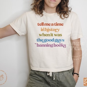 Op de afbeelding: Een wit T-shirt met een regenboogkleurige afbeelding met de tekst "tell me a time in history when it was the good guys banning books".