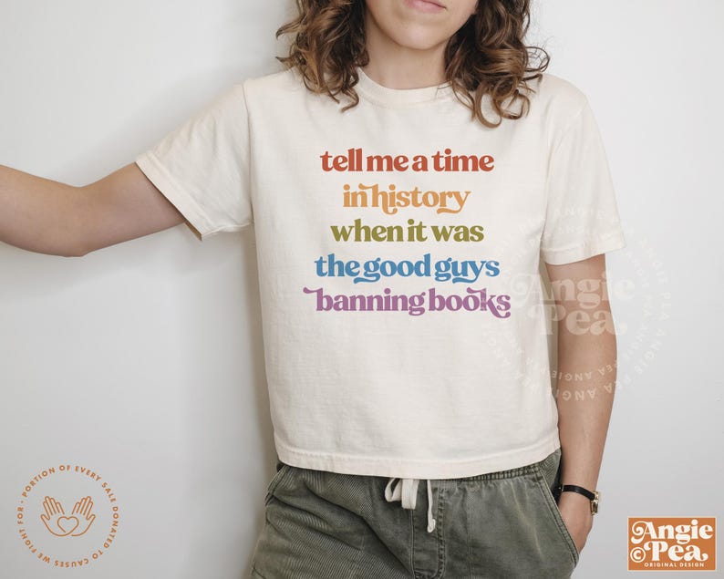 Op de afbeelding: Een wit T-shirt met een regenboogkleurige afbeelding met de tekst "tell me a time in history when it was the good guys banning books".