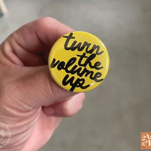 May include: A yellow button with the phrase "turn the volume up" in black script. The button is round and has a glossy finish. The button is held in a hand.