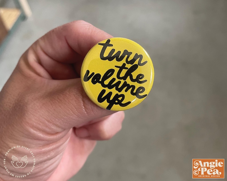 May include: A yellow button with the phrase "turn the volume up" in black script. The button is round and has a glossy finish. The button is held in a hand.