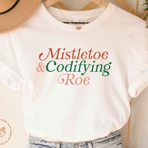 May include: White t-shirt with the text "Mistletoe & Codifying Roe" in green, pink, and brown lettering.
