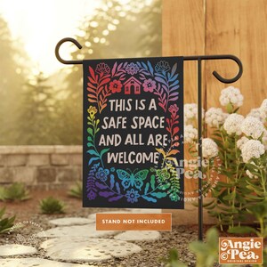 May include: A black garden flag with a rainbow floral design and the text "This is a safe space and all are welcome".