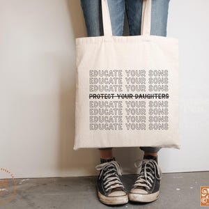 Op de afbeelding: Een witte tote bag met de tekst "Educate your sons" die meerdere keren wordt herhaald, met de zin "Protect your daughters" in vetgedrukte letters in het midden van de tas.
