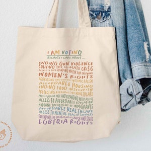 May include: A beige tote bag with the text "I AM VOTING BECAUSE I CARE ABOUT..." and a list of social and political issues in rainbow colors. The issues include ending gun violence, solving the climate crisis, women's rights, affordable housing, and LGBTQIA rights.