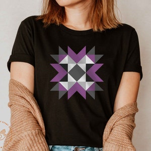 May include: A black t-shirt with a purple, gray, and white geometric design. The design is a quilt block pattern.
