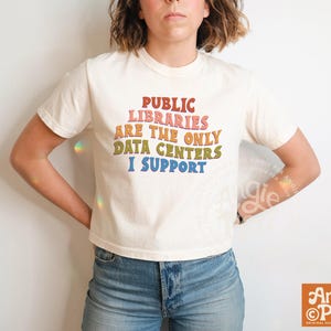 May include: Cream-colored t-shirt with the text "PUBLIC LIBRARIES ARE THE ONLY DATA CENTERS I SUPPORT" in colorful retro lettering. The shirt is a short-sleeved crew neck.