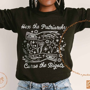May include: Black sweatshirt with a white graphic design featuring a hand with an eye, a tooth, and stars. The text "Hex the Patriarchy" and "Curse the Bigots" is printed on the sweatshirt.