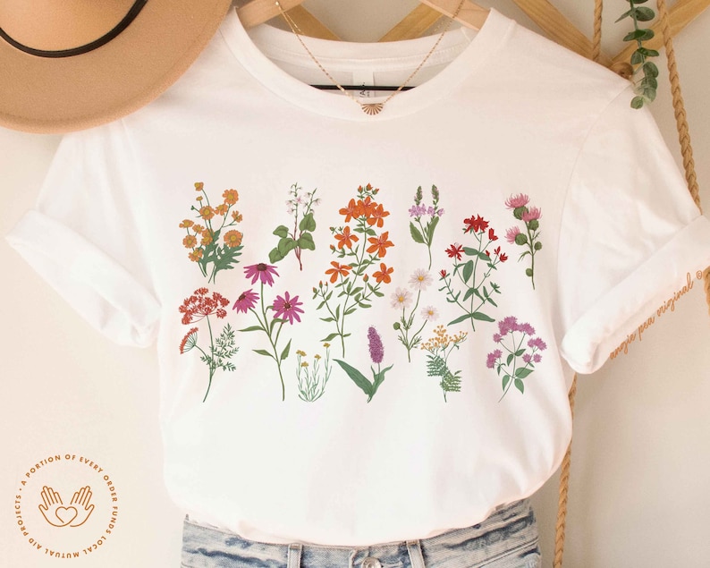 May include: White t-shirt with a floral print featuring a variety of flowers in shades of orange, pink, red, and yellow. The shirt has a round neckline and short sleeves. The text "A portion of every order funds local mutual aid projects." is printed on the bottom left corner of the shirt.