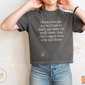 May include: Dark gray t-shirt with the text "i feared that one day we'd have to watch men burn the world down, i just didn't expect them to be such losers" in white. The shirt is paired with blue jeans.