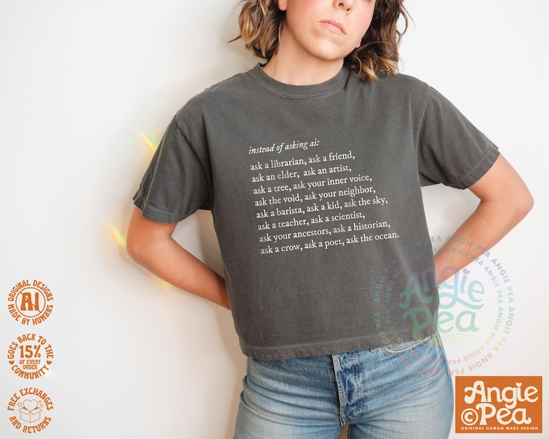 May include: Dark gray t-shirt with white text that reads "instead of asking ai: ask a librarian, ask a friend...ask the ocean." The shirt is paired with blue jeans. The design is by Angie Pea.