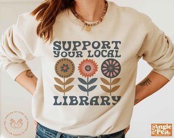 May include: A beige sweatshirt with the text "Support Your Local Library" in a retro font. The text is surrounded by three stylized flowers in shades of brown, pink, and blue.