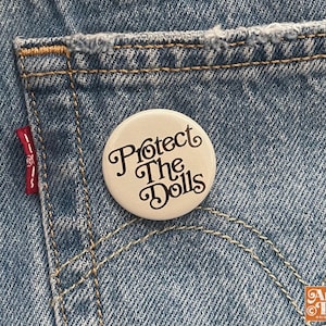 May include: A white button pin with the text "Protect The Dolls" in a decorative font. The pin is set against a backdrop of blue denim jeans, with visible stitching and a red tag.
