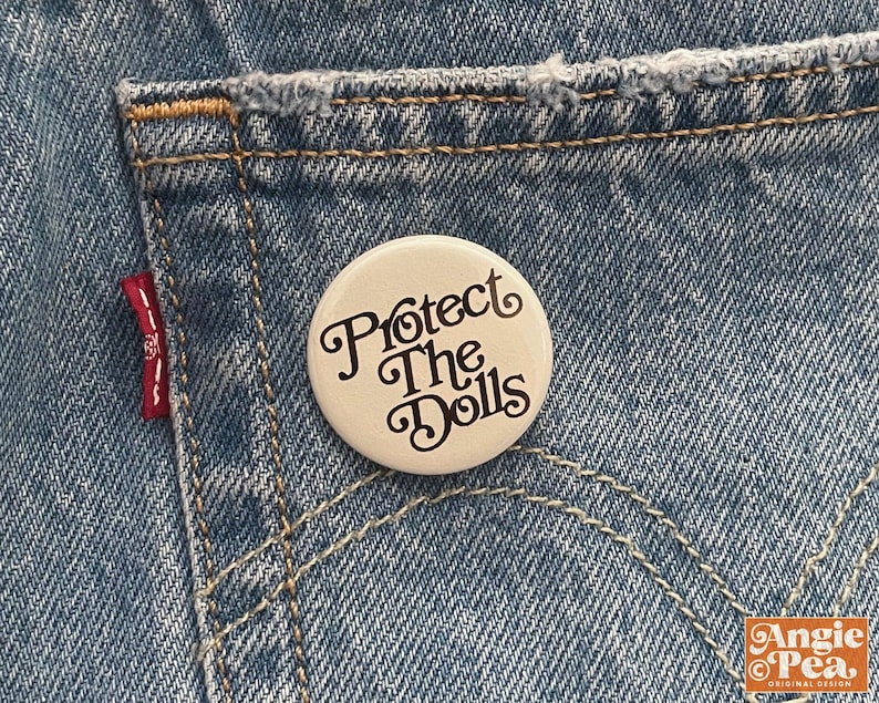 May include: A white button pin with the text "Protect The Dolls" in a decorative font. The pin is set against a backdrop of blue denim jeans, with visible stitching and a red tag.