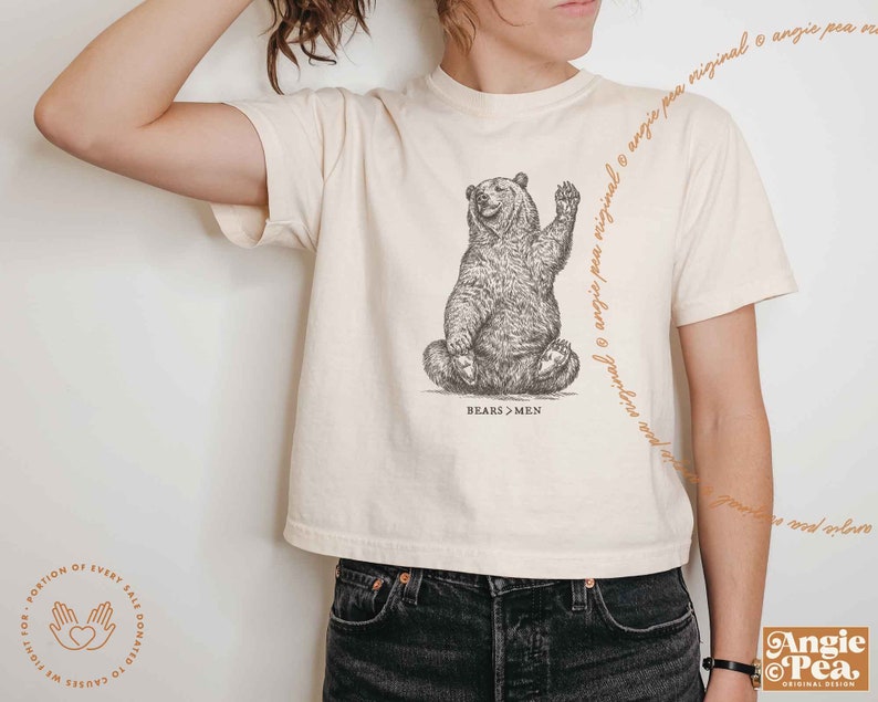 K&ouml;nnte beinhalten: Ein wei&szlig;es, bauchfreies T-Shirt mit einer Schwarzwei&szlig;-Illustration eines sitzenden und winkenden B&auml;ren. Der Text "BEARS > MEN" ist unter dem B&auml;ren aufgedruckt.