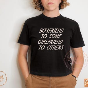 May include: Black crew neck t-shirt with the text "BOYFRIEND TO SOME GIRLFRIEND TO OTHERS" in white. The shirt is worn with brown pants. The Angie Pea logo is visible on the lower right.