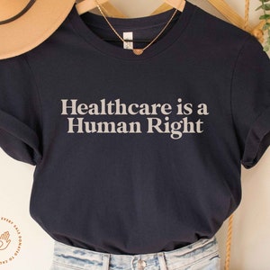 May include: A navy blue t-shirt with the text "Healthcare is a Human Right" printed in white on the front. The shirt is folded and displayed on a wooden hanger.