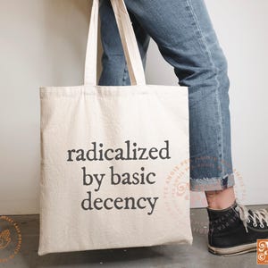 May include: A cream-colored tote bag with the phrase "radicalized by basic decency" printed in black. The bag has two handles and is being held by someone wearing blue jeans and black sneakers.