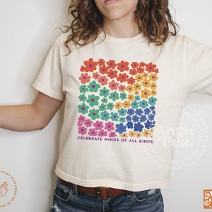 May include: Cream-colored t-shirt featuring a square floral design in rainbow colors. The shirt has the text "CELEBRATE MINDS OF ALL KINDS" printed below the flowers. The shirt is a casual, everyday wear item.