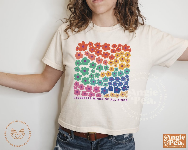 May include: Cream-colored t-shirt featuring a square floral design in rainbow colors. The shirt has the text "CELEBRATE MINDS OF ALL KINDS" printed below the flowers. The shirt is a casual, everyday wear item.