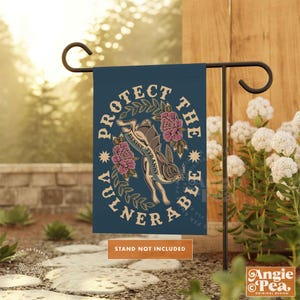 May include: A dark teal garden flag with the words "PROTECT THE VULNERABLE" encircling a rabbit illustration. The rabbit holds a banner reading "RESIST TYRANNY". Pink flowers and stars accent the design. The flag is displayed on a black metal garden stake.