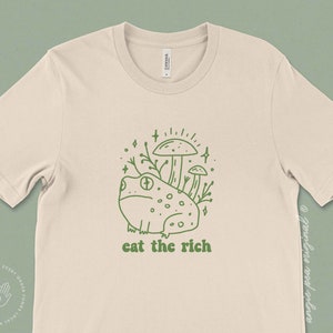 May include: A beige t-shirt featuring a green line drawing of a frog sitting on top of mushrooms with the text "eat the rich" underneath.