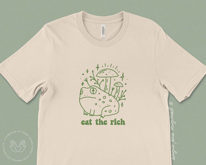 May include: A beige t-shirt featuring a green line drawing of a frog sitting on top of mushrooms with the text "eat the rich" underneath.