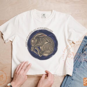 May include: Cream-colored t-shirt featuring a circular illustration of a sleeping hare within a dark blue circle. The text "NONE OF US ALIENS. ALL OF US EARTHLINGS" is printed around the image. The shirt is paired with blue jeans.