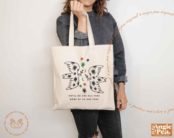 Könnte beinhalten: Eine beige Tragetasche mit einer schwarz-weißen Illustration von zwei Vögeln mit grünen und roten Akzenten. Der Text "Until we are all free, none of us are free" ist auf die Tasche gedruckt.