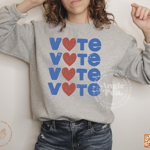 以下が含まれることがあります： 青い文字で「VOTE」という単語が4回繰り返され、各単語の間に赤いハートが描かれたグレーのトレーナーです。