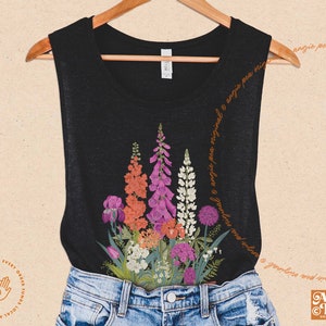 May include: Black sleeveless top featuring a vibrant floral design with purple, orange, and white flowers. The top is paired with denim shorts. The text "Angie Pea Original Design" is visible.