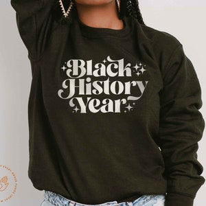 K&ouml;nnte beinhalten: Dunkel olivgr&uuml;nes Sweatshirt mit dem Text "Black History Year" in wei&szlig;er Retro-Schrift. Der Text ist von Sternen umgeben. Das Sweatshirt hat einen Rundhalsausschnitt.