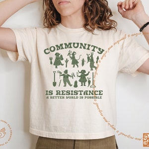 May include: A white t-shirt with a green graphic design that says "COMMUNITY IS RESISTANCE A BETTER WORLD IS POSSIBLE". The graphic features a group of people gardening.