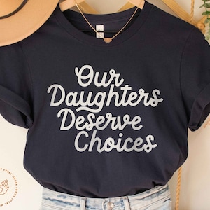 May include: A navy blue t-shirt with the text "Our Daughters Deserve Choices" printed in silver on the front.