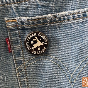 May include: A round black pin with white text and a rabbit graphic is pinned to a denim pocket. The pin reads "DEFY, DERAIL, DISRUPT, FASCISM." The denim is light blue with visible stitching and a red Levi's tag.