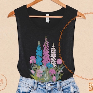 May include: A black tank top with a floral design featuring pink, blue, and purple flowers. The flowers are arranged in a cluster and appear to be a variety of wildflowers. The tank top is worn over a pair of blue jeans.