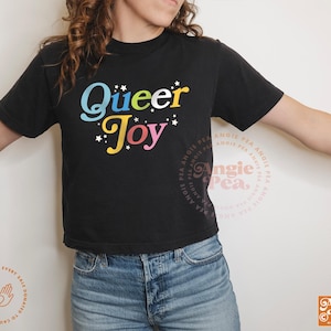 May include: Black t-shirt with the words "Queer Joy" in colorful, playful lettering. The design includes stars and a retro font. The shirt is a crew neck and is paired with blue jeans.