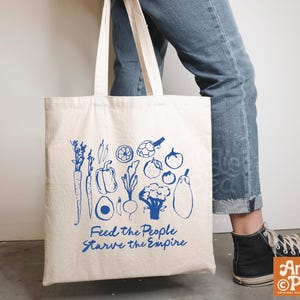 May include: A cream-colored canvas tote bag featuring a blue graphic print of various vegetables including carrots, peppers, and broccoli. Below the produce, the text reads, Feed the People Starve the Empire in a blue, handwritten-style font.